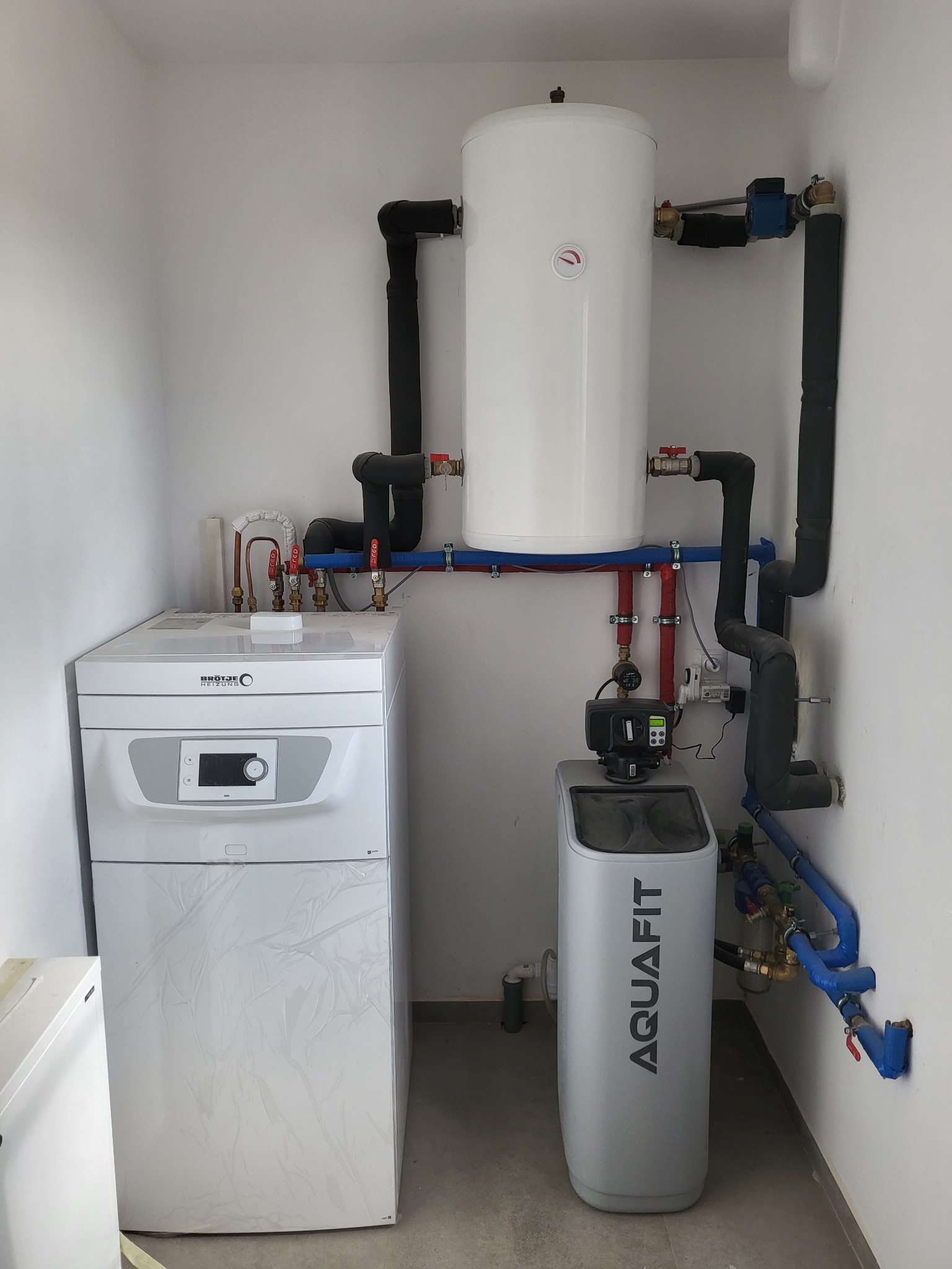 Biała kotłownia z kotłem Brötje, zasobnikiem ciepłej wody i filtrem Aquafit, rury w izolacji, widoczne elementy instalacji hydraulicznej.