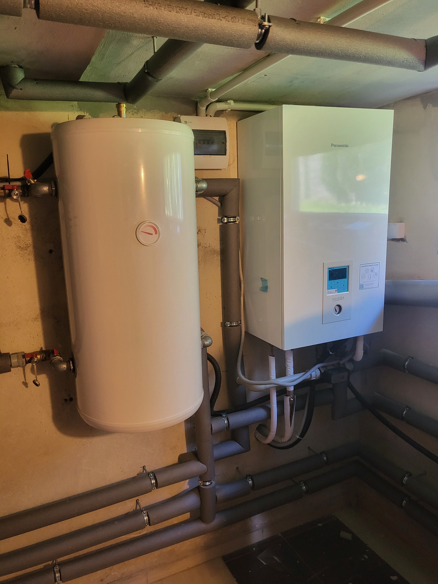 Biały zasobnik ciepłej wody z termometrem i biała jednostka wewnętrzna z wyświetlaczem, połączone szarymi rurami w piwnicy lub pomieszczeniu technicznym.