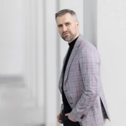 Mężczyzna w kraciastej marynarce i czarnym golfie, oparty o białą kolumnę w Czechowicach-Dziedzicach, pozujący do zdjęcia.