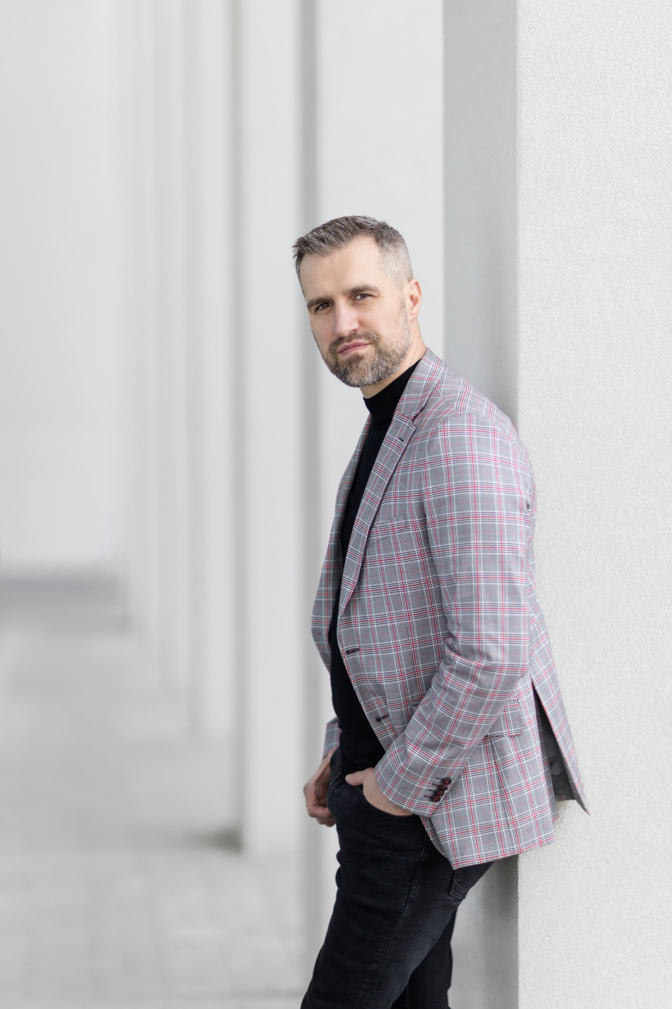 Mężczyzna w kraciastej marynarce i czarnym golfie, oparty o białą kolumnę w Czechowicach-Dziedzicach, pozujący do zdjęcia.