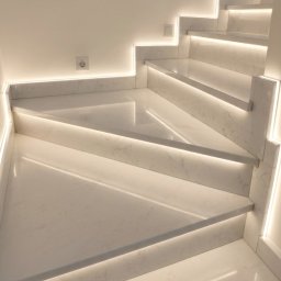 Płytki Max - Białe schody z marmuru carrara, podświetlone listwami LED, tworzące nowoczesny efekt wizualny. Widoczne gniazdko elektryczne na ścianie.