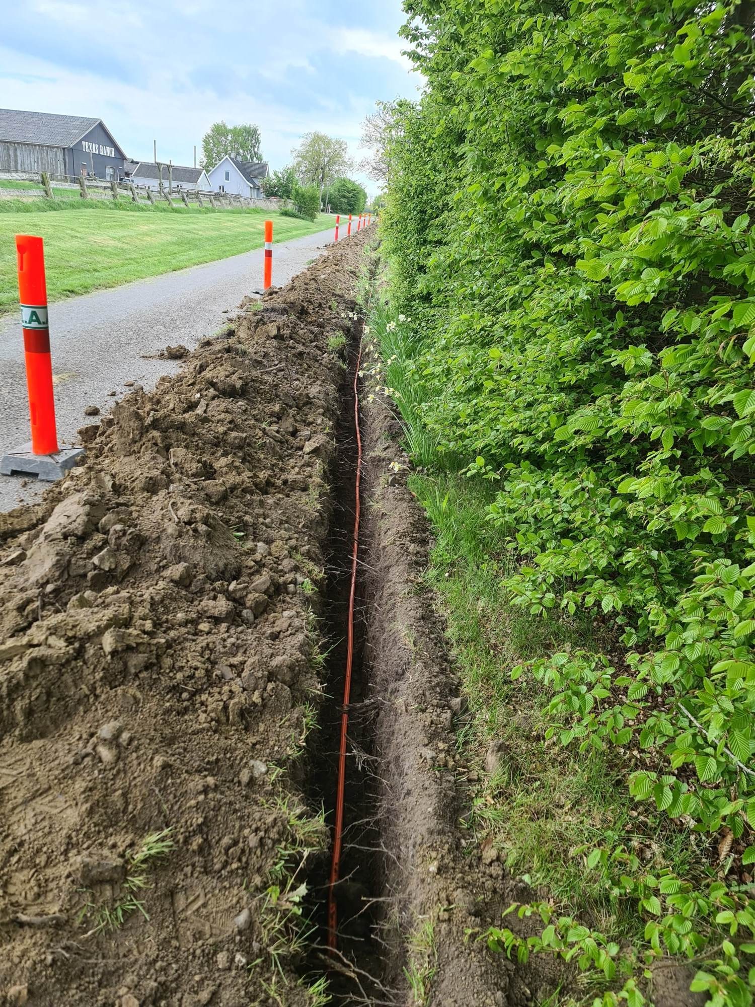 Wąski wykop wzdłuż ścieżki, zabezpieczony pomarańczowymi słupkami, z widocznym pomarańczowym przewodem wewnątrz, sąsiadujący z zielonym żywopłotem i budynkami w tle.