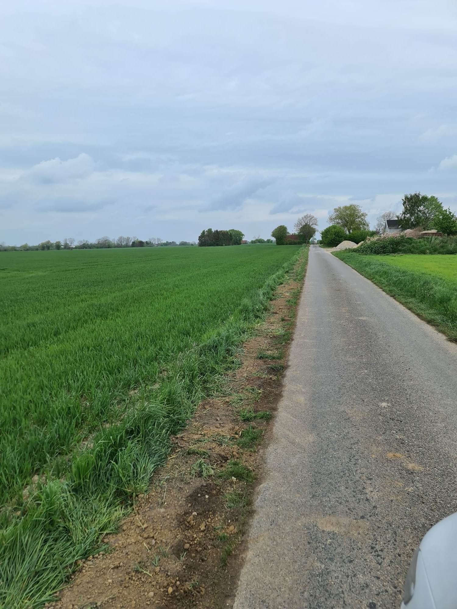 Asfaltowa droga biegnąca przez wiejski krajobraz, z polami po lewej stronie i luźną stertą ziemi po prawej, pod pochmurnym niebem.