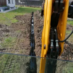 Wąski wykop wypełniony wodą, wykonany na trawniku obok domu z panelami słonecznymi, widoczny z perspektywy żółtej koparki.