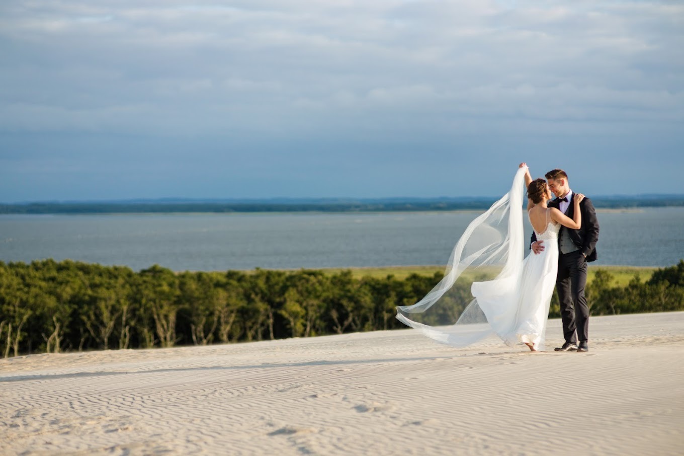Para młoda na piaszczystej wydmie, panna młoda w zwiewnej sukni i welonie, w tle widok na jezioro i las.