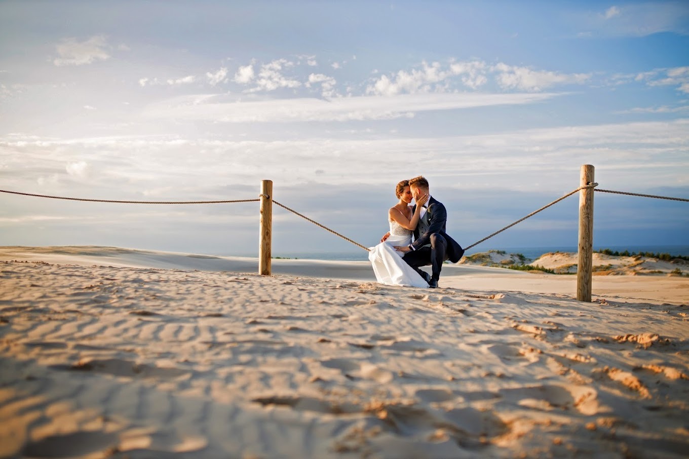 Para młoda siedzi na linie między drewnianymi słupkami na piaszczystej wydmie, błękitne niebo z chmurami w tle, delikatne światło zachodzącego słońca.
