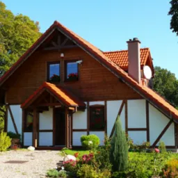 Dwukondygnacyjny dom szkieletowy z czerwonym dachem, białymi ścianami z drewnianą kratownicą i anteną satelitarną, otoczony zielenią i kwiatami w słoneczny dzień.