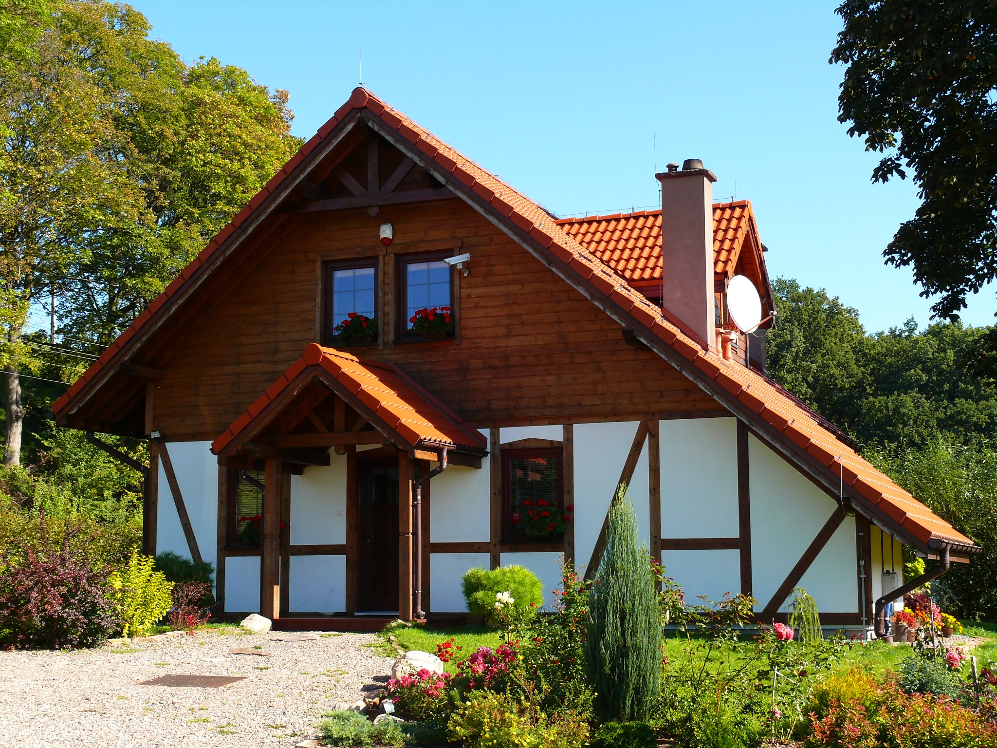 Dwukondygnacyjny dom szkieletowy z czerwonym dachem, białymi ścianami z drewnianą kratownicą i anteną satelitarną, otoczony zielenią i kwiatami w słoneczny dzień.