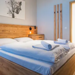 Nowoczesna sypialnia z drewnianym łóżkiem, szafkami nocnymi i panelem za łóżkiem; na ścianie grafika z motywem drzewa i ludzkiej twarzy, w tle widoczne drewniane drzwi z czarnymi paskami.