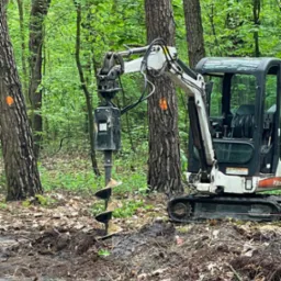 Mini koparka z wiertłem świdrowym wiercąca otwór w ziemi na zalesionym terenie, z drzewami oznaczonymi pomarańczową farbą.
