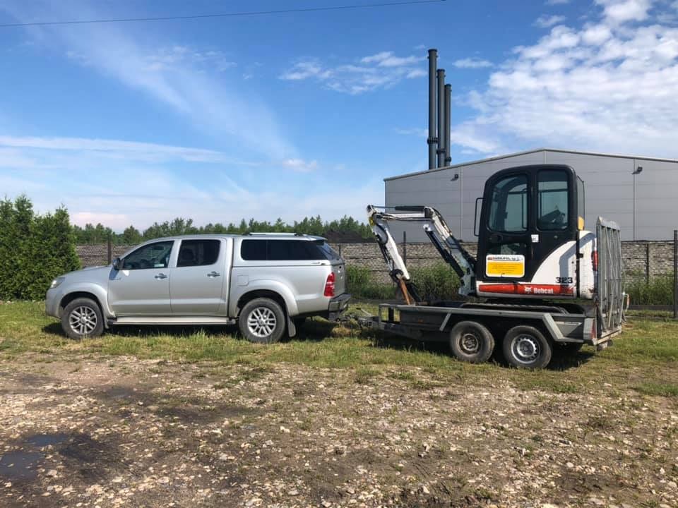 Srebrny pickup Toyota Hilux z przyczepą przewożącą minikoparkę Bobcat na tle budynku przemysłowego i błękitnego nieba z chmurami.