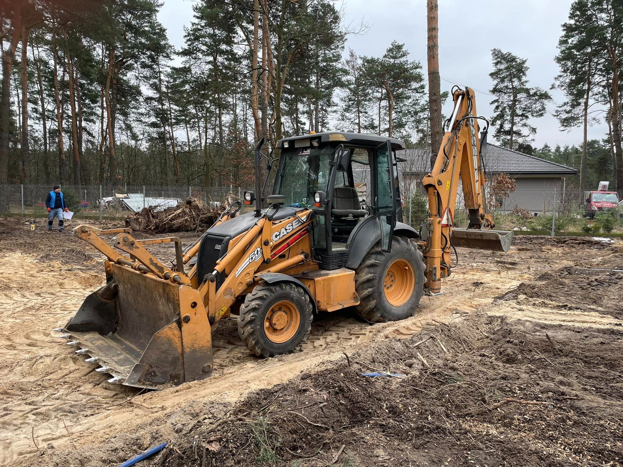 Żółto-czarna koparko-ładowarka Case 580ST na piaszczystym placu budowy, w tle drzewa i częściowo widoczny dom. Na dalszym planie pracownik w niebieskiej kurtce i czerwony samochód dostawczy.