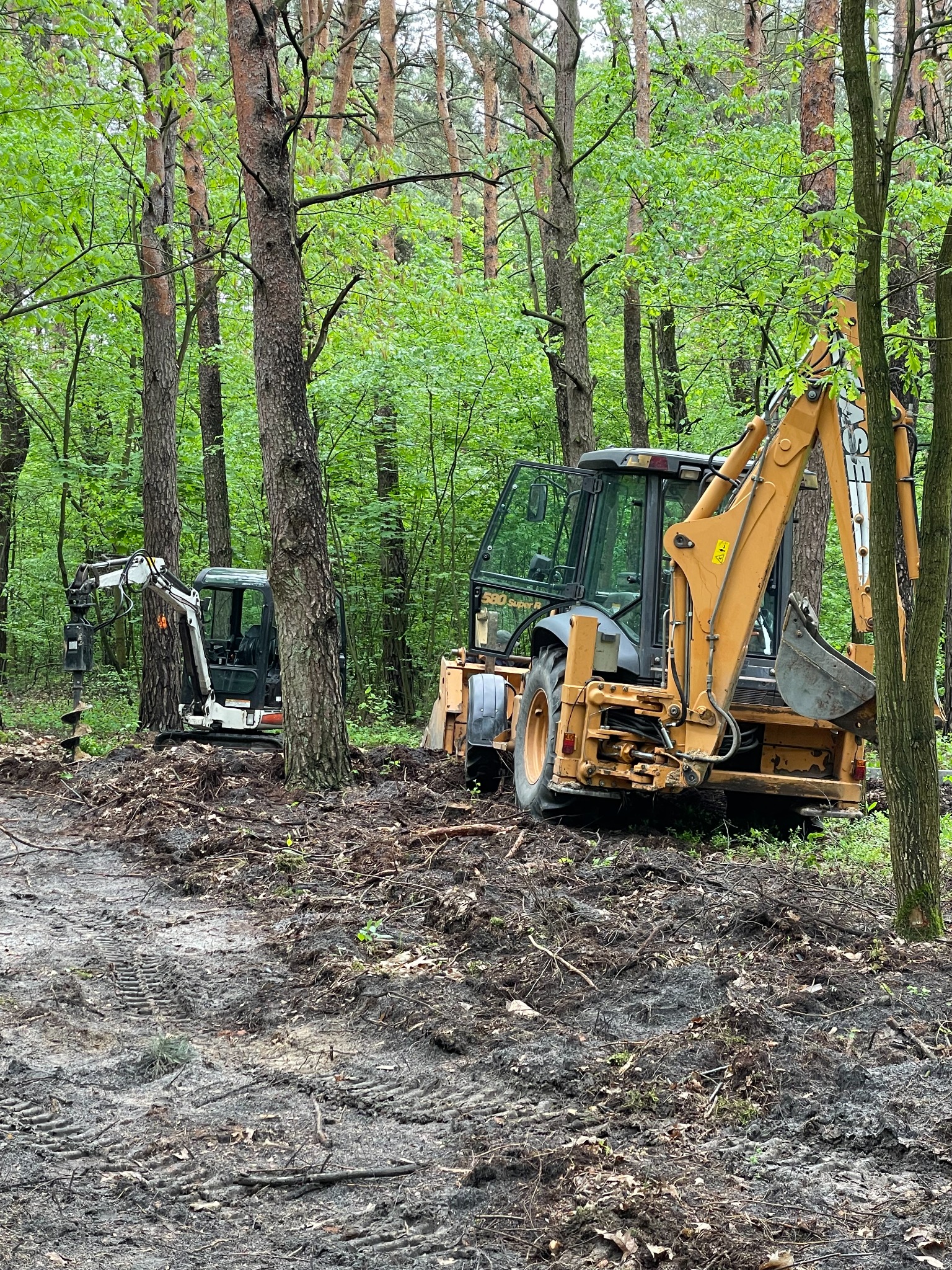 Dwie maszyny budowlane, koparka i mini koparka z wiertnicą, pracują na zalesionym terenie, przygotowując teren pod budowę; widoczne ślady opon na błotnistym gruncie.