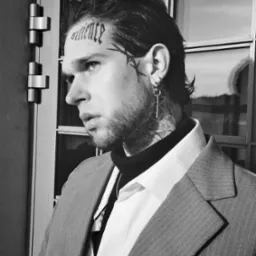 Czarno-białe zdjęcie portretowe mężczyzny z tatuażami na twarzy i szyi, ubranego w garnitur, stojącego blisko okna.