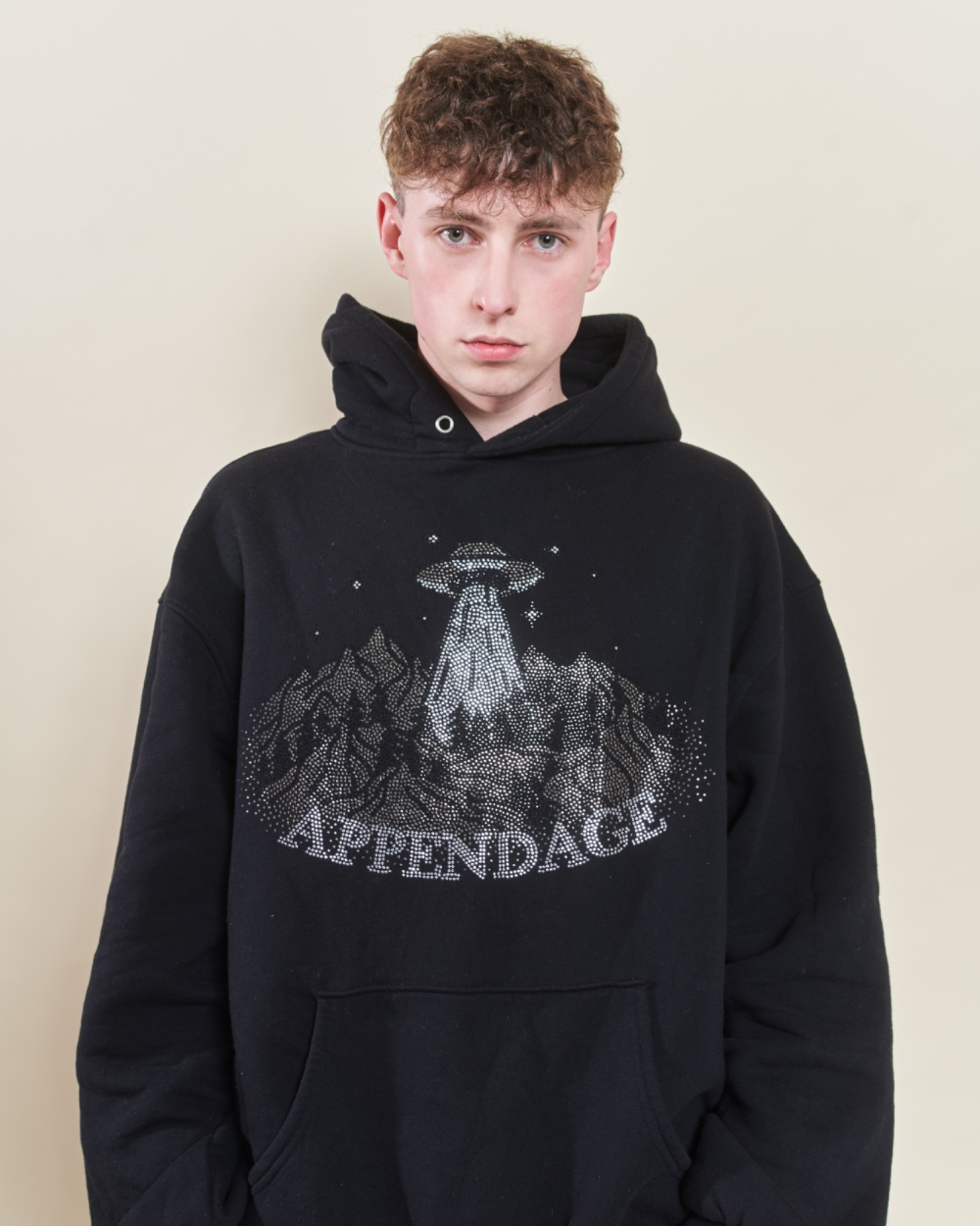 Młody mężczyzna pozuje w czarnej bluzie z kapturem, ozdobionej błyszczącym motywem UFO nad górami i napisem 'APPENDAGE'.