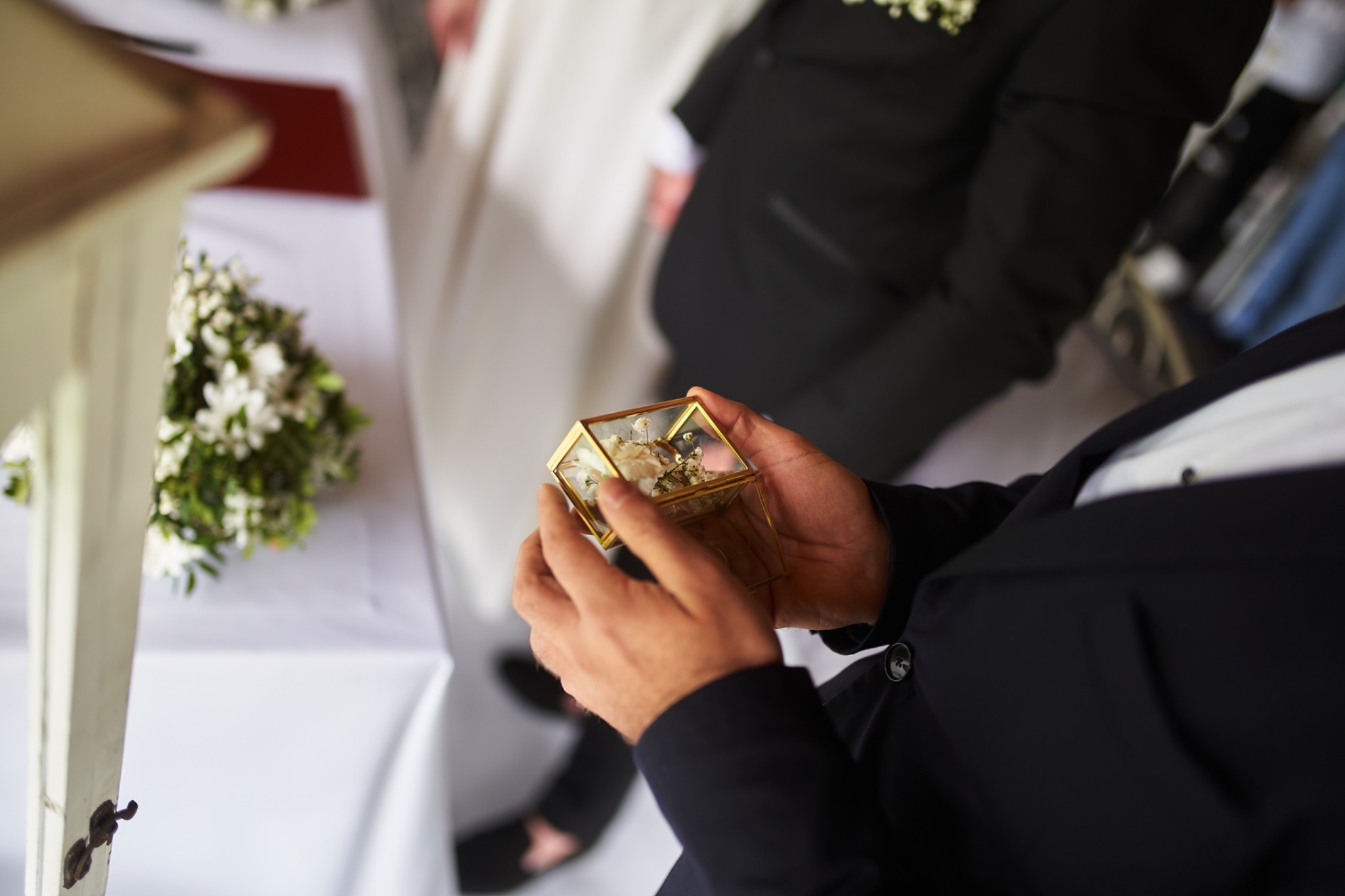 Mężczyzna w garniturze trzyma w dłoniach szklane pudełko z kwiatami i pierścionkami wewnątrz, w tle bukiet białych kwiatów i fragment sukni ślubnej.