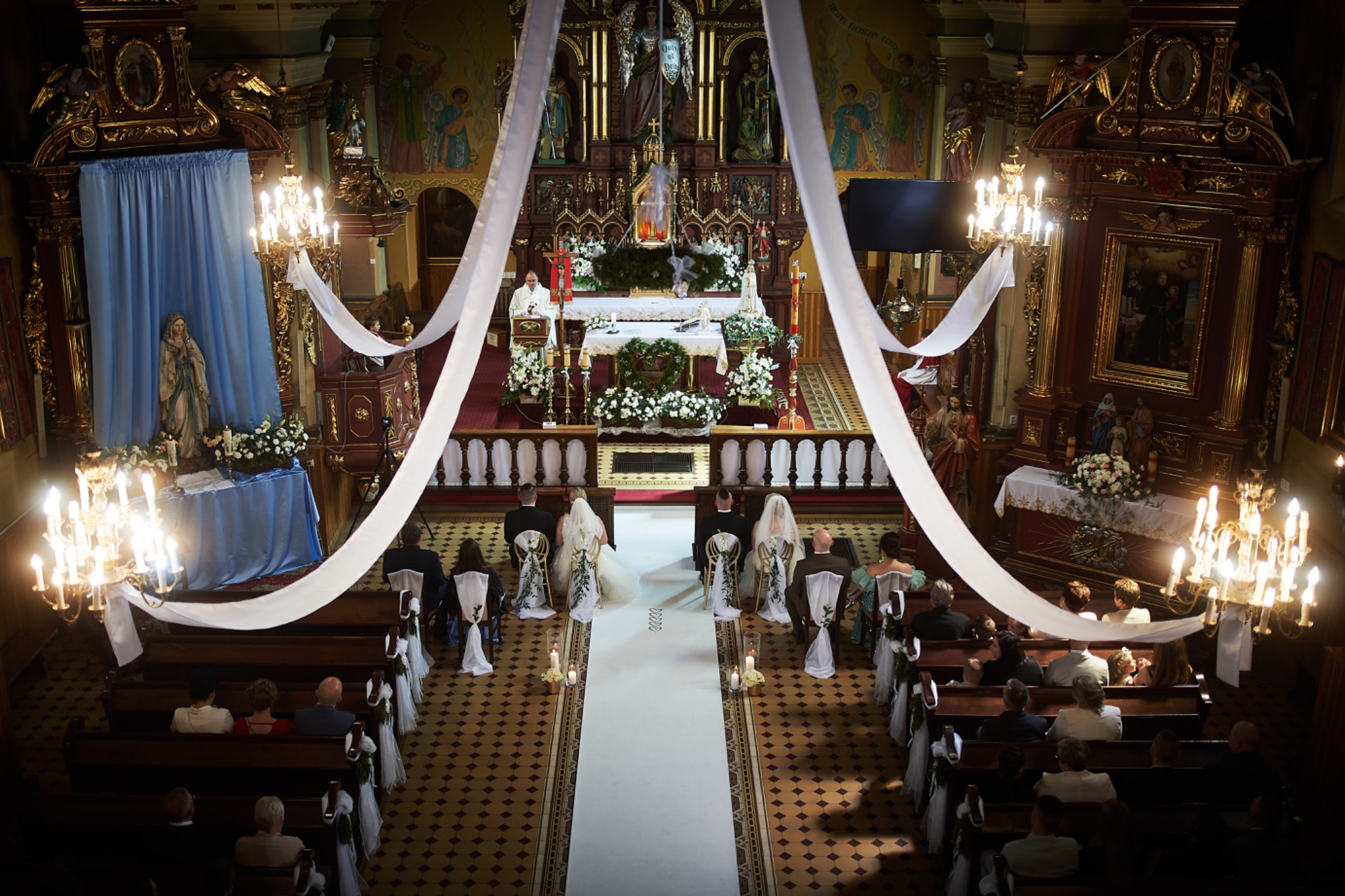 Widok z góry na parę młodą siedzącą w kościele podczas ceremonii ślubnej, z białym dywanem prowadzącym do ołtarza, ozdobionym kwiatami i świecami, oraz białymi wstążkami dekorującymi ławki.