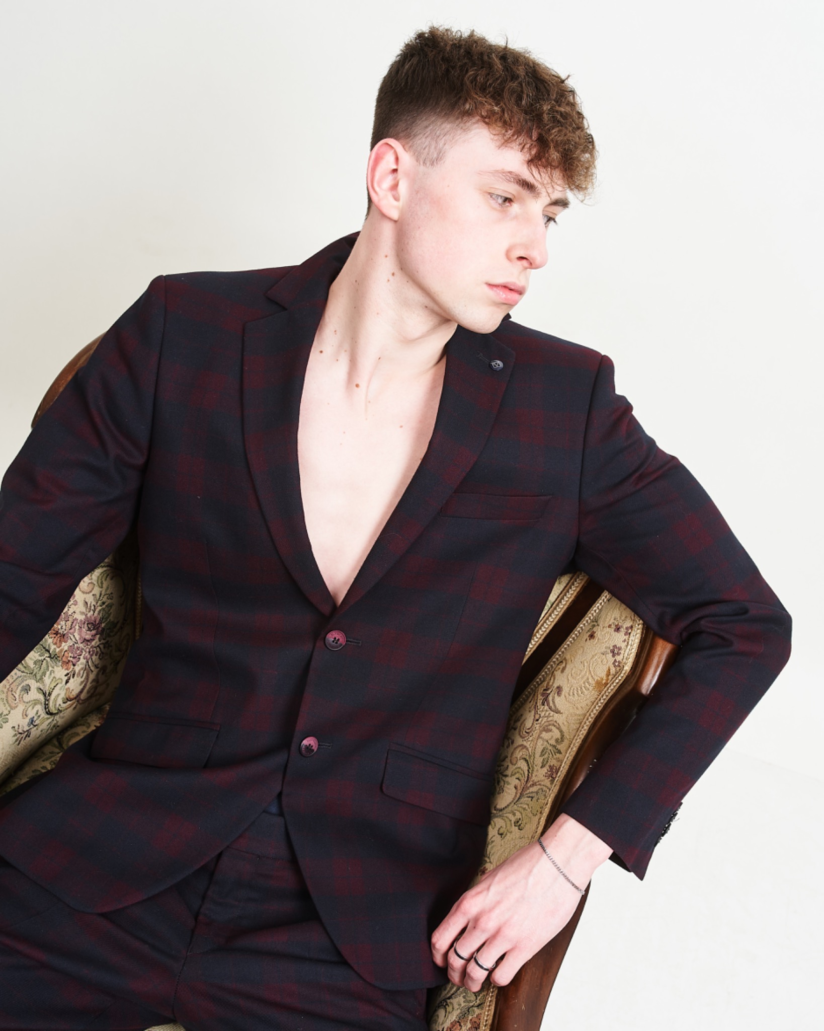 Młody mężczyzna w rozpiętym, bordowo-granatowym garniturze w kratę, siedzący w fotelu z kwiecistą tapicerką, z bransoletką na nadgarstku i dwoma pierścionkami.