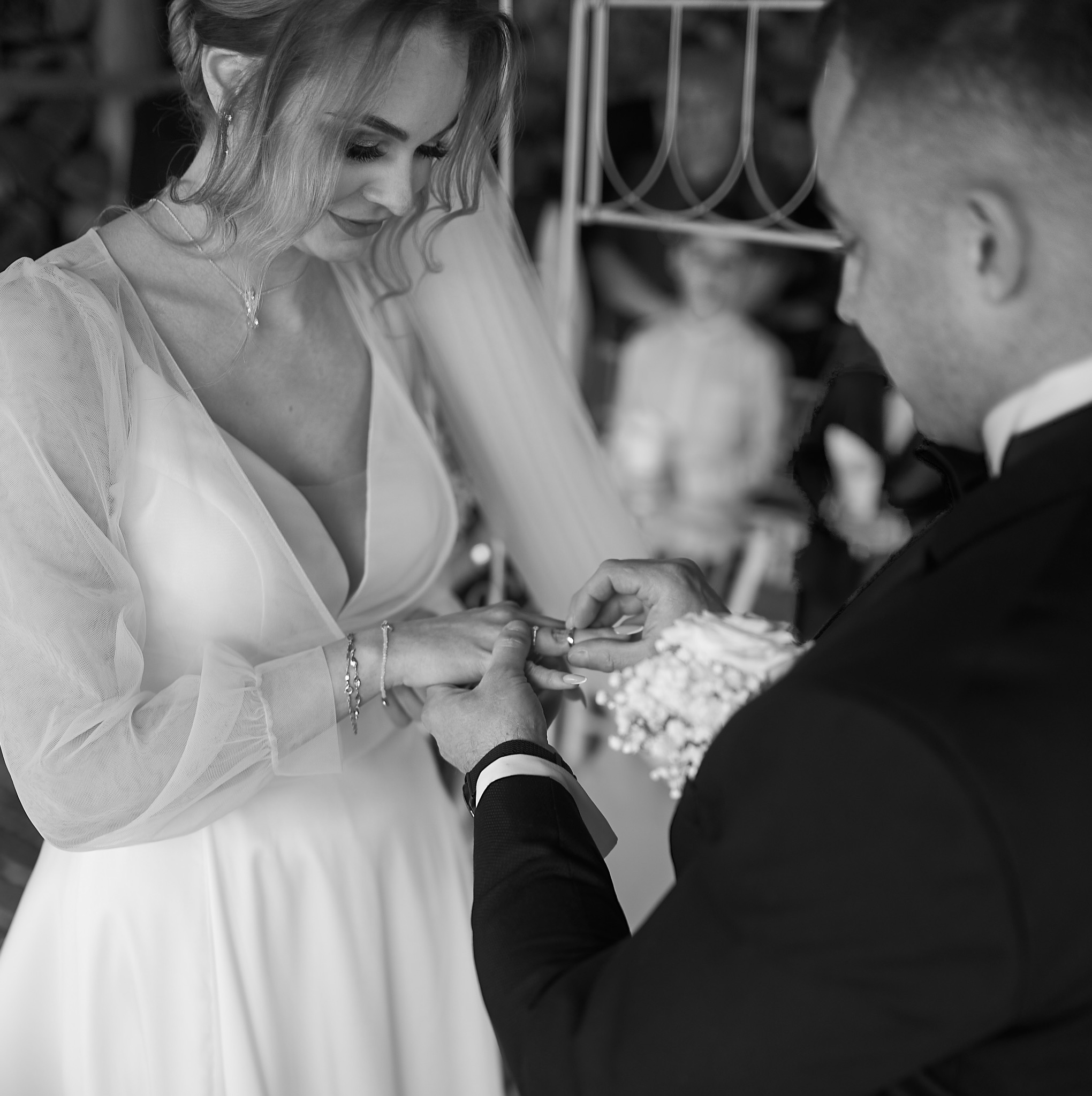 Czarno-białe zdjęcie przedstawia zbliżenie dłoni panny młodej, na której palec pan młody zakłada obrączkę. Panna młoda ma na sobie suknię z delikatnego materiału i biżuterię. W tle widoczny zarys...