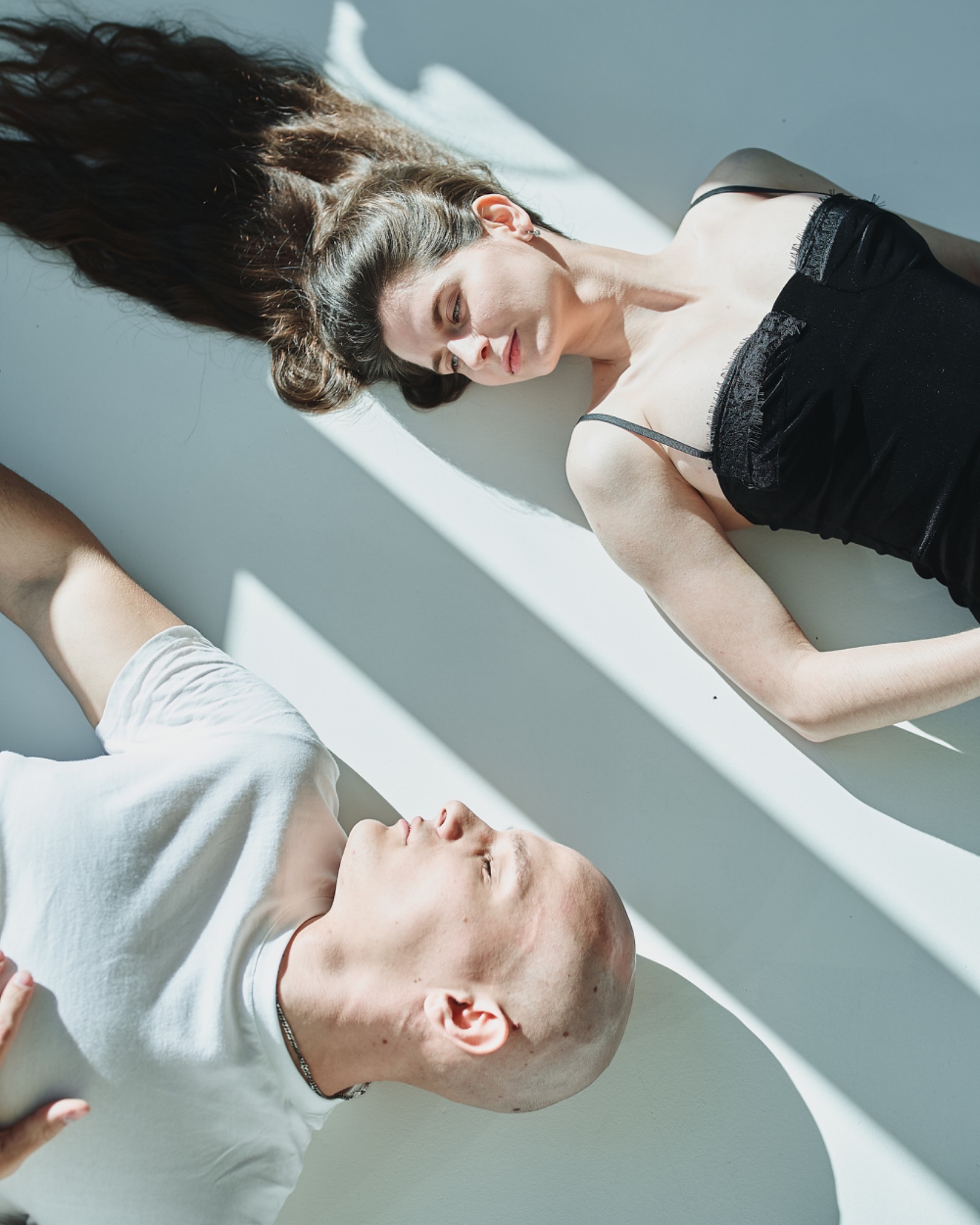 Kobieta w czarnej bieliźnie i mężczyzna w białej koszulce leżą na białym tle w mocnym, kontrastowym świetle słonecznym, tworząc cienie.