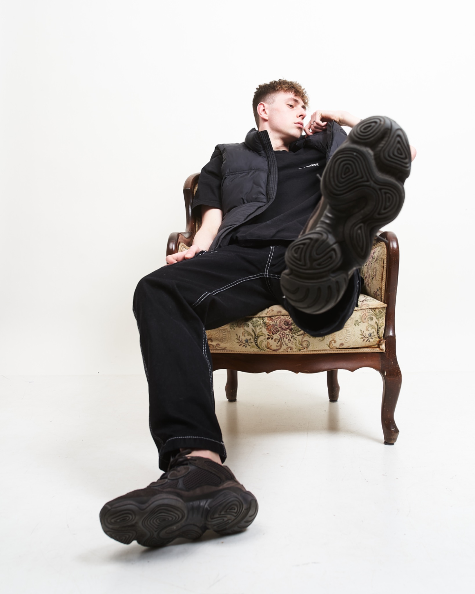 Młody mężczyzna w czarnym ubraniu relaksuje się w fotelu, eksponując podeszwę buta w kierunku aparatu.