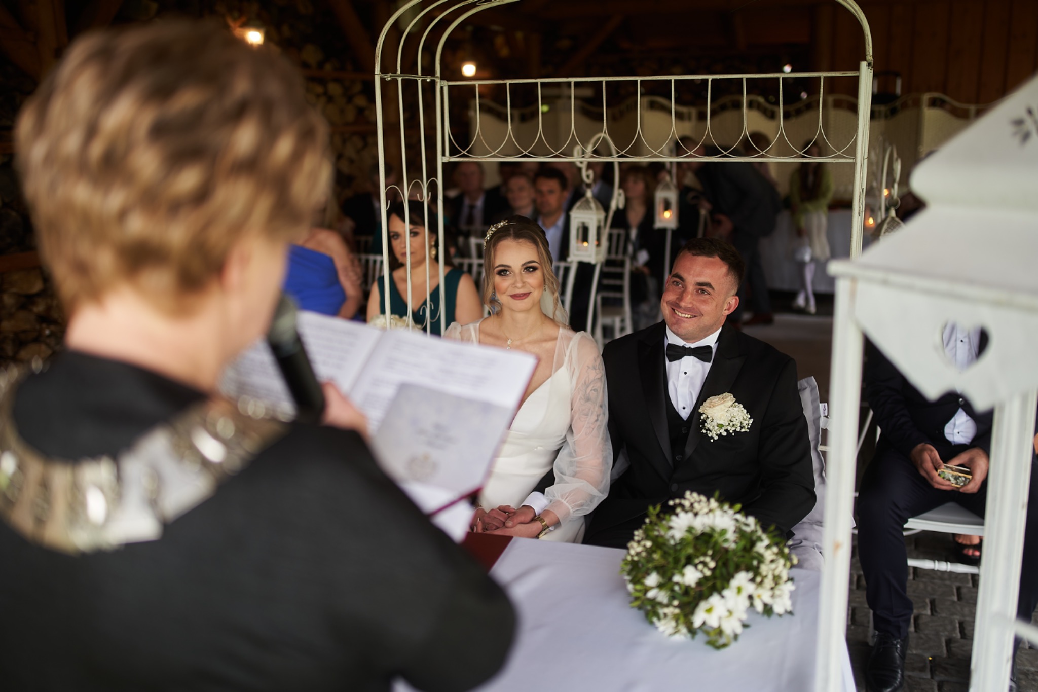 Para młoda podczas ceremonii ślubnej, siedząca pod białym, metalowym baldachimem, z bukietem na stole. W tle widoczni goście, a na pierwszym planie osoba przemawiająca do mikrofonu.