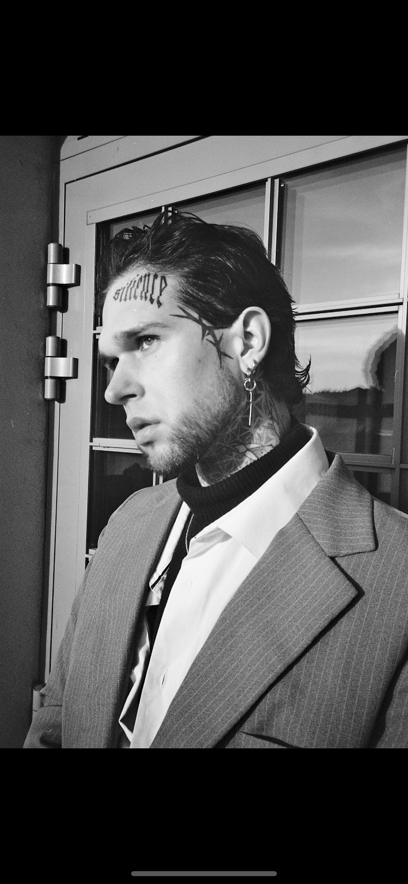 Czarno-białe zdjęcie portretowe mężczyzny z tatuażami na twarzy i szyi, ubranego w garnitur, stojącego blisko okna.
