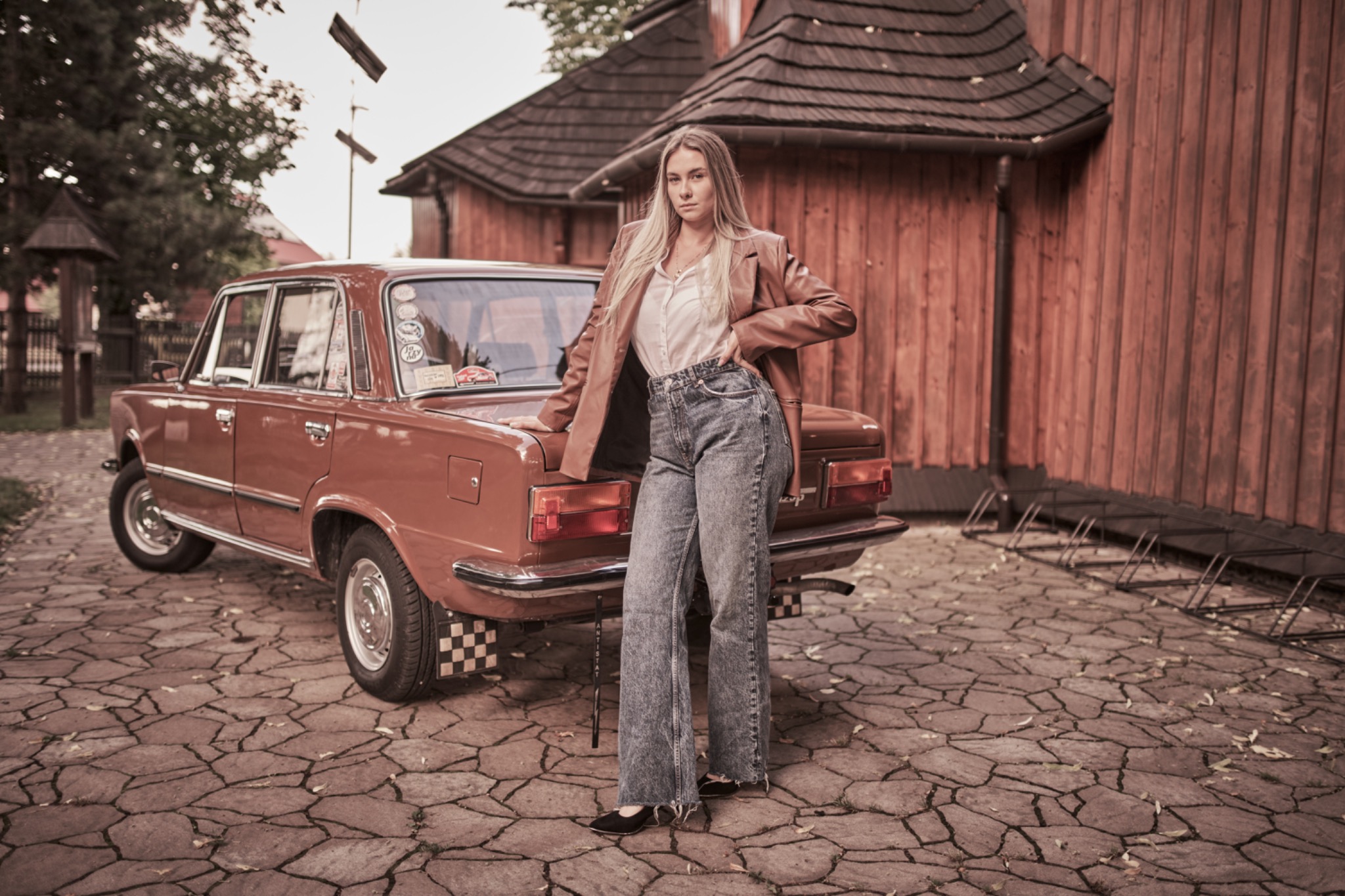 Blondwłosa kobieta w dżinsach i brązowej marynarce pozuje oparta o pomarańczowy samochód na tle drewnianego budynku o spadzistym dachu