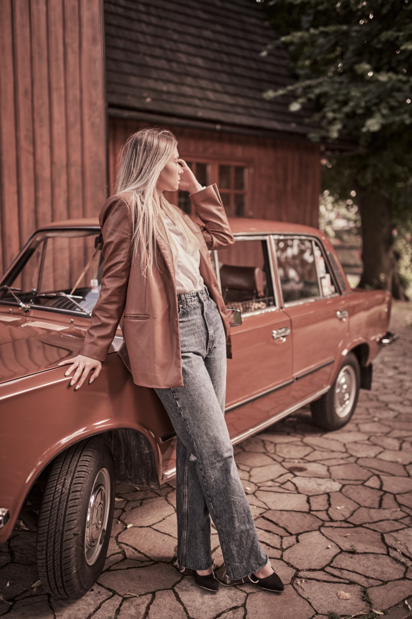 Kobieta w brązowej marynarce i dżinsach opiera się o czerwony samochód na tle drewnianego budynku i drzewa.