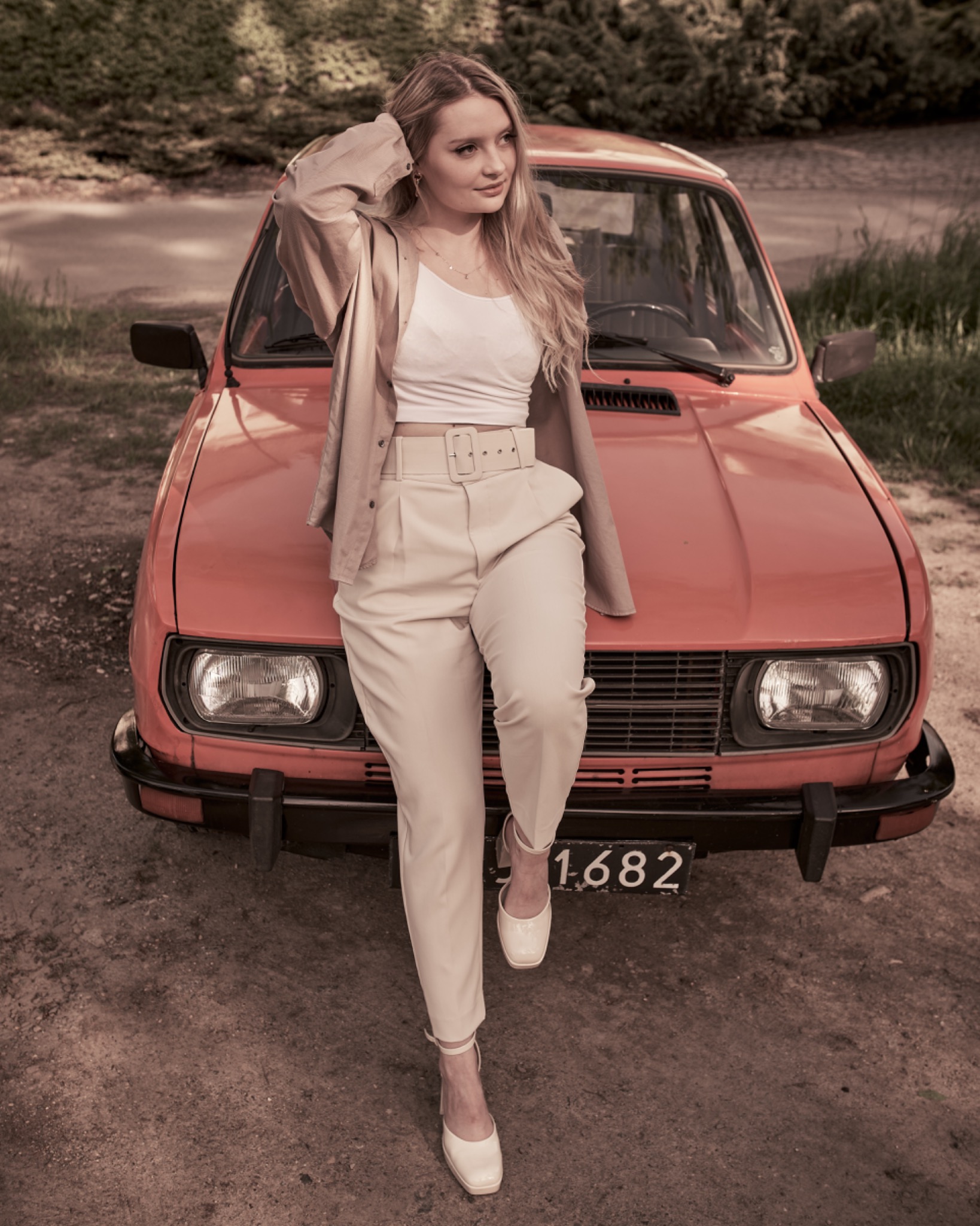 Kobieta w beżowym garniturze opiera się o pomarańczowy samochód na polnej drodze, z ręką we włosach i spojrzeniem w bok.