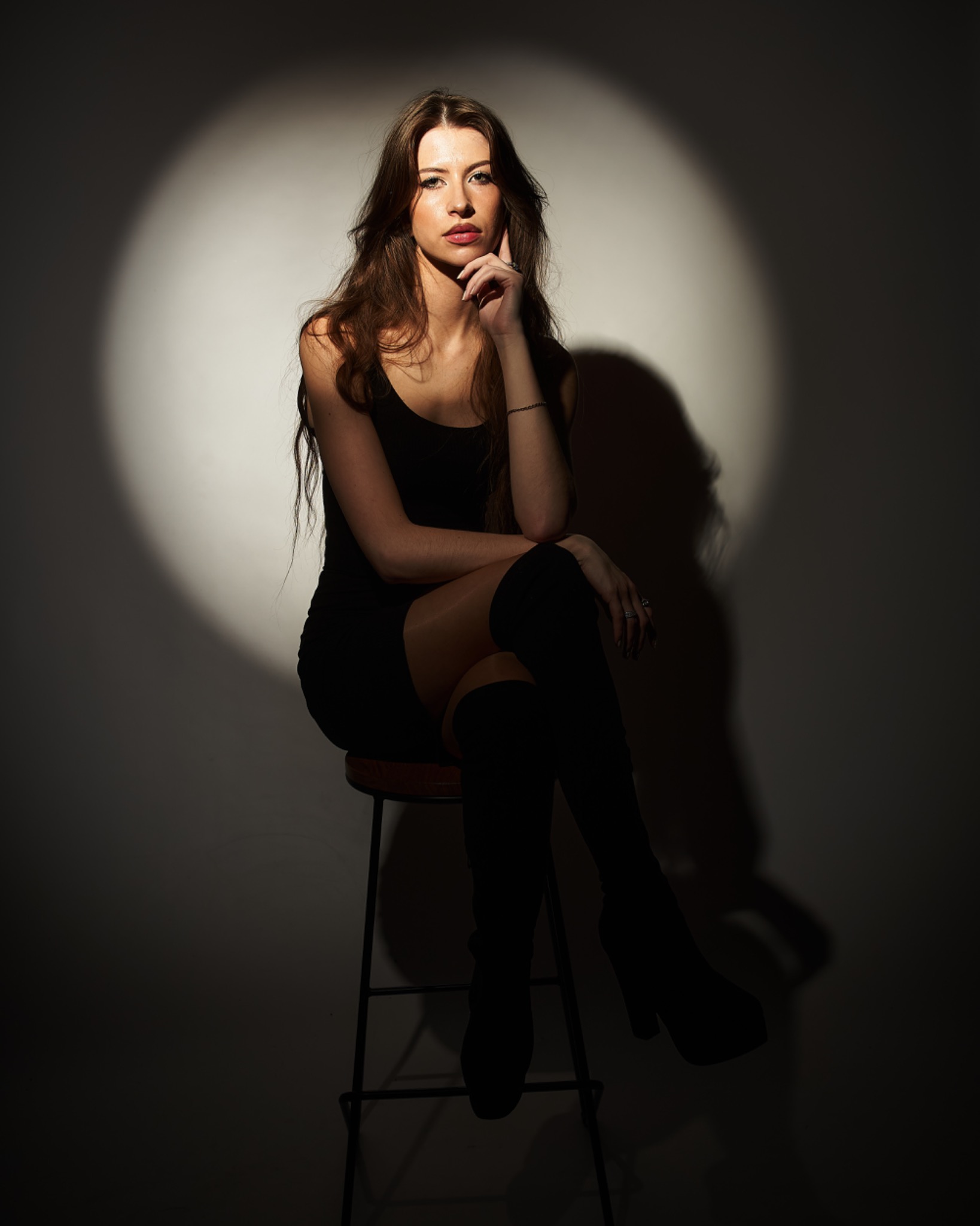 Portret kobiety w czarnej sukience i kozakach, siedzącej na stołku w studiu w Oświęcimiu, oświetlonej punktowym światłem, zamyślona poza.