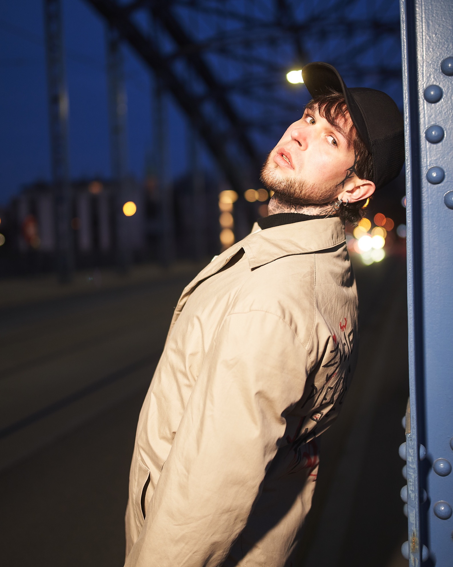 Młody mężczyzna w beżowym płaszczu z nadrukiem na rękawie, oparty o niebieską konstrukcję mostu w wieczornym świetle, z widocznymi tatuażami na szyi i twarzy.