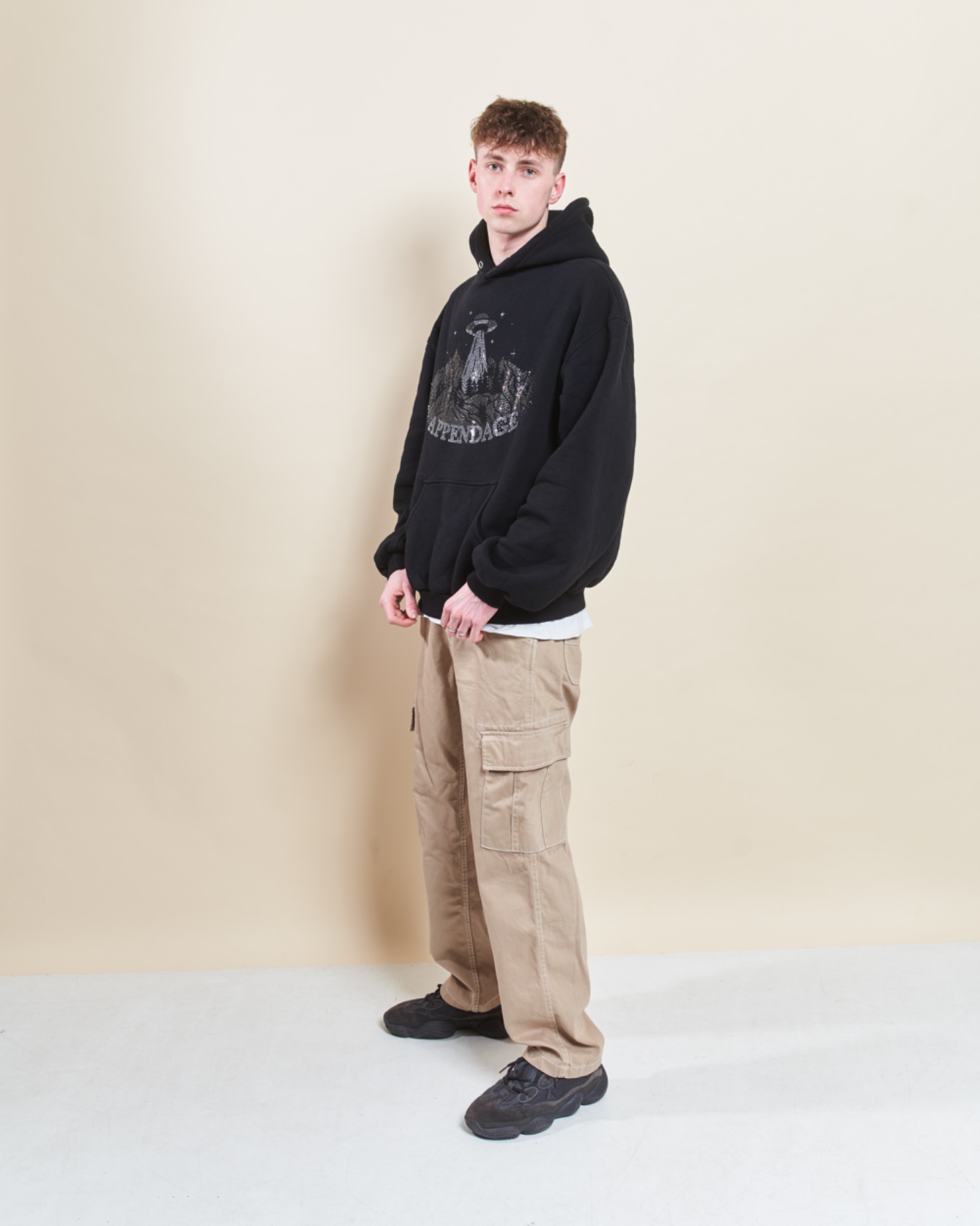 Młody mężczyzna pozuje w czarnej bluzie z kapturem z nadrukiem UFO i beżowych spodniach cargo, stojąc na jasnym tle.