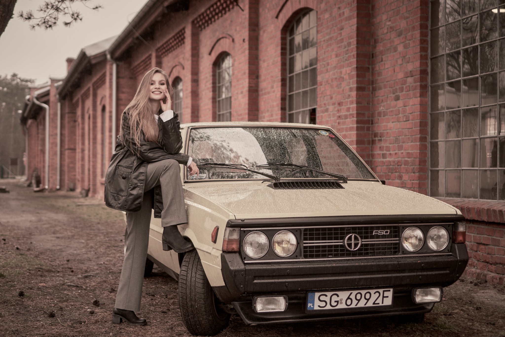 Uśmiechnięta kobieta w płaszczu i spodniach pozuje oparta o maskę klasycznego samochodu FSO Polonez na tle ceglanego budynku w deszczowy dzień.