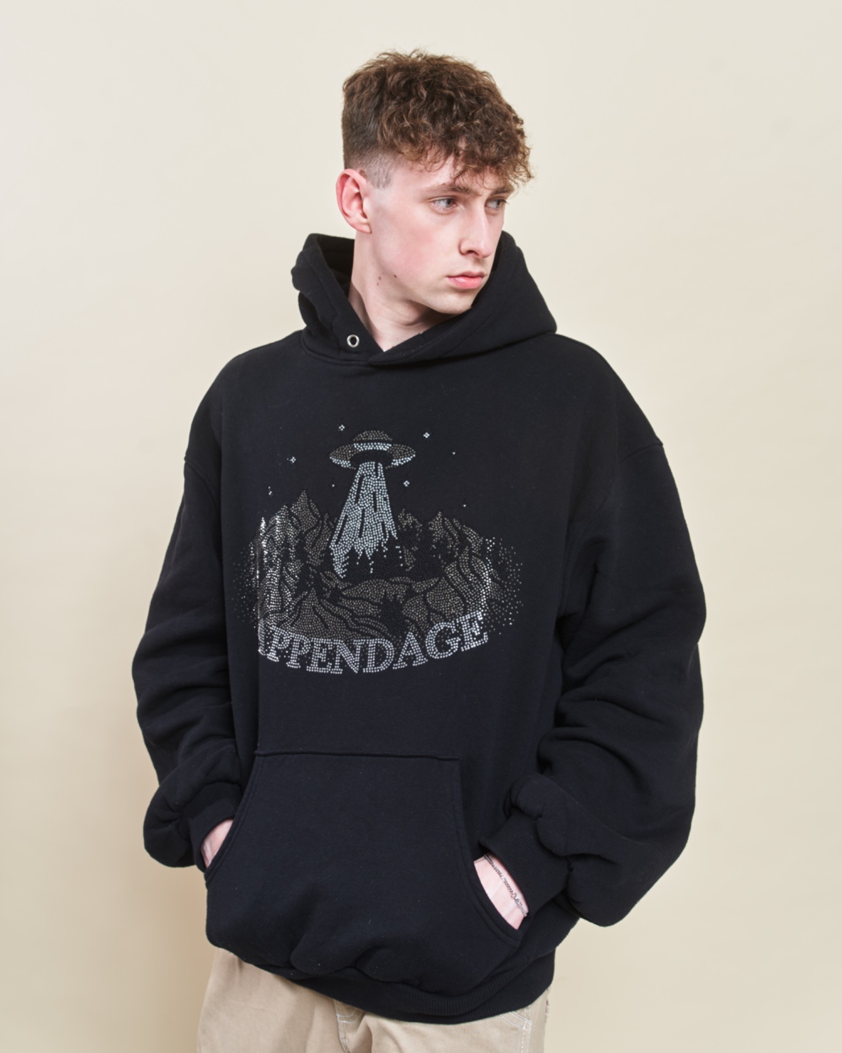Młody mężczyzna pozuje w czarnej bluzie z kapturem, na której widnieje aplikacja z motywem UFO nad górami, wykonana z błyszczących dżetów, z napisem 'APPENDAGE' poniżej. Tło jest jednolite, kremowe.