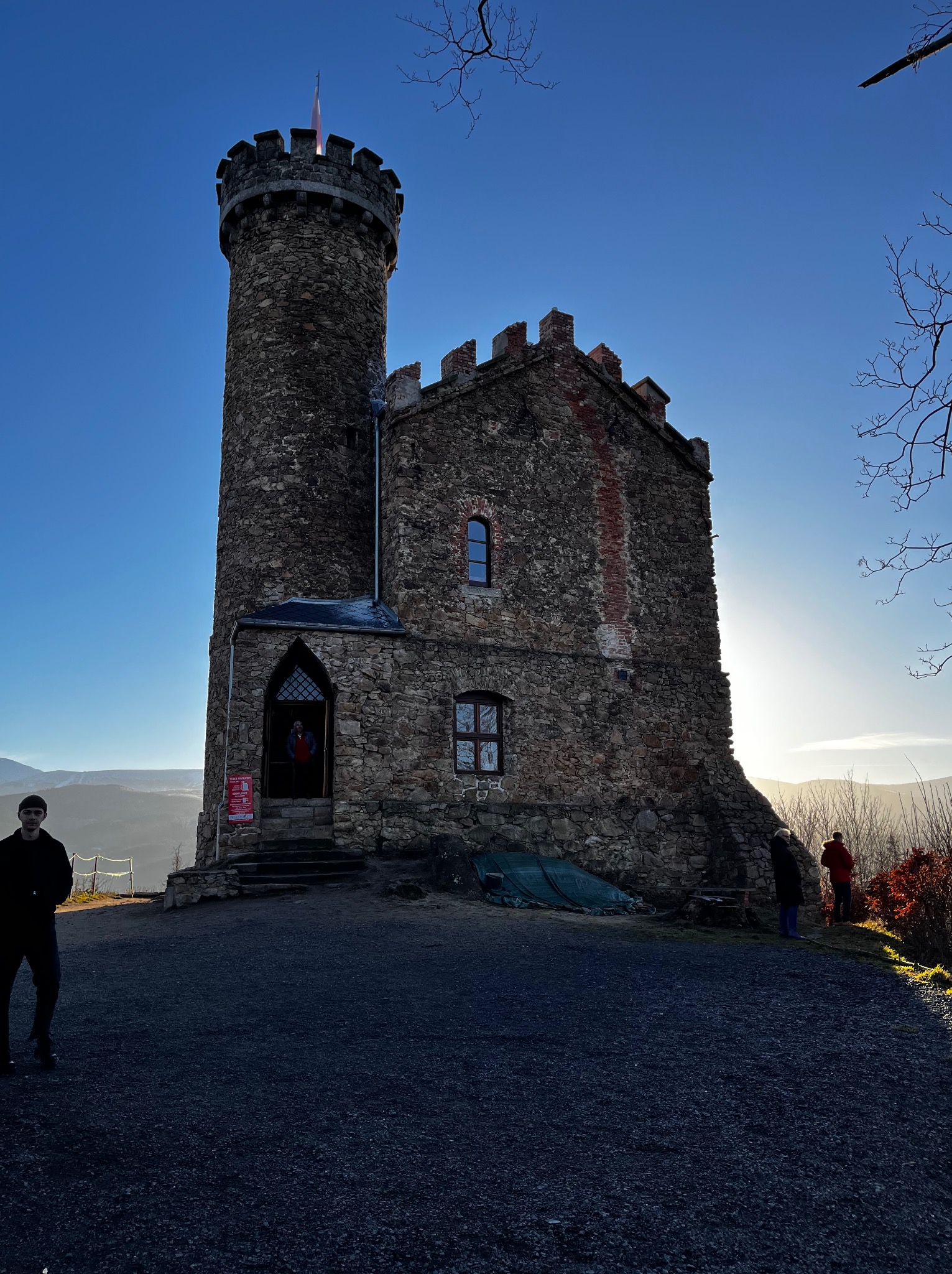 Kamienna wieża widokowa z blankami i małym oknem, w tle góry i błękitne niebo. Na pierwszym planie osoba i fragment ścieżki.