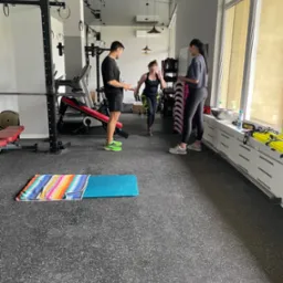 Kobieta w trakcie treningu w siłowni, nadzorowana przez dwóch trenerów, z widocznym sprzętem do ćwiczeń i matą na podłodze.