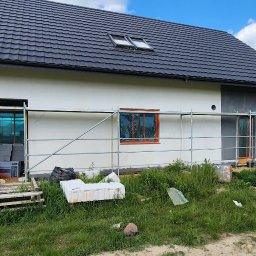 Morozov BUD - Dom jednorodzinny w trakcie ocieplania elewacji. Widoczne rusztowania, okna zabezpieczone folią, częściowo otynkowana ściana. W tle pole rzepaku i błękitne niebo.