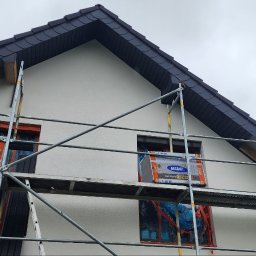 Morozov BUD - Prace na rusztowaniu przy ociepleniu domu. Widoczny fragment dachu z ciemnej dachówki. Okna zabezpieczone folią. Elementy rusztowania. Praca w toku, dzień.