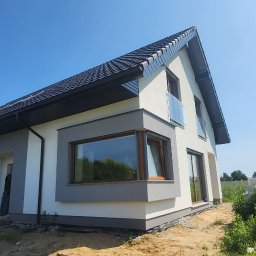 Morozov BUD - Nowoczesny dom jednorodzinny z ciemną dachówką, szarymi elementami elewacji i dużymi oknami, widziany z perspektywy trawnika. Częściowo widoczne materiały budowlane.