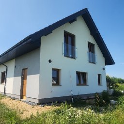 Morozov BUD - Nowoczesny, dwukondygnacyjny dom z ciemnym dachem i białymi ścianami, z widocznymi oknami z drewnianą stolarką i szklanymi balustradami balkonowymi, otoczony zielenią.