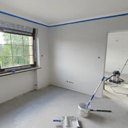 Morozov BUD - Pokój po malowaniu ścian na szaro, z niebieską taśmą malarską przy suficie i oknach. Widoczne narzędzia malarskie i osoba w trakcie prac wykończeniowych.