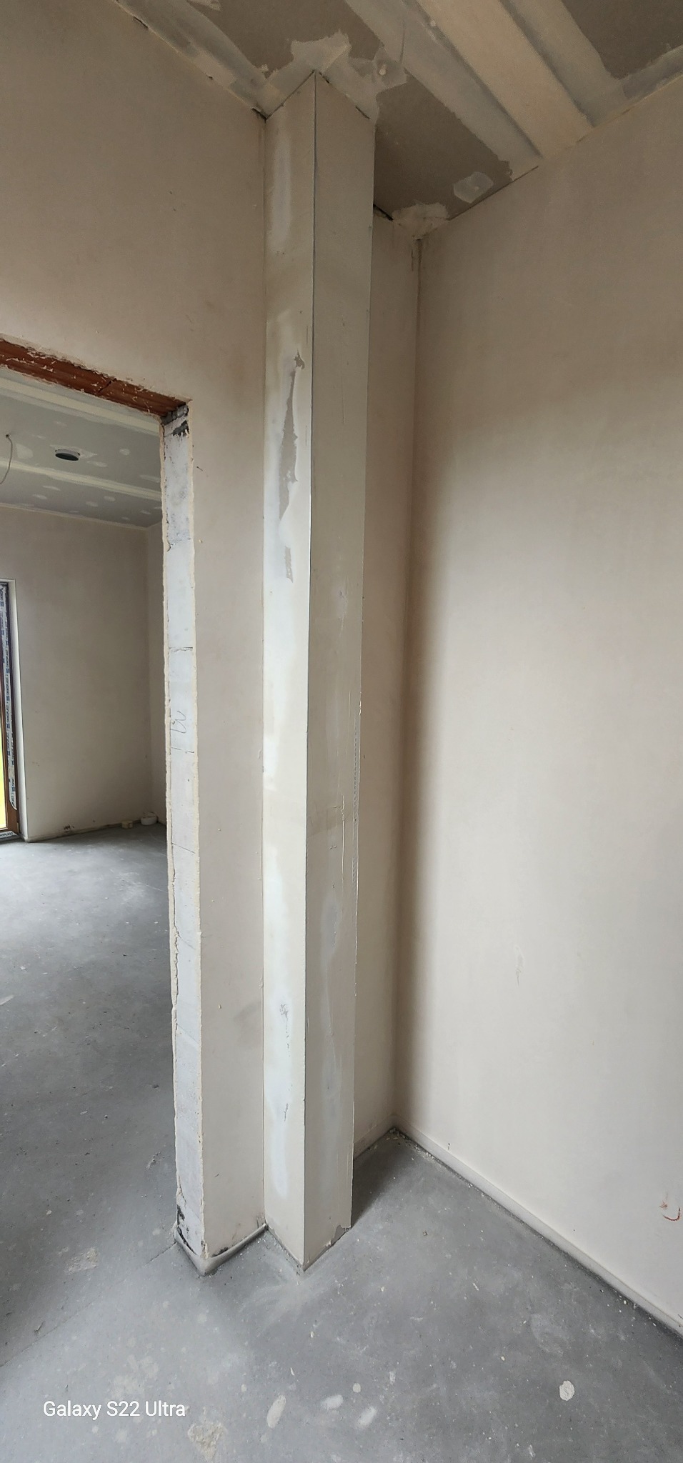 Wnętrze w trakcie prac wykończeniowych: ściany częściowo wyszpachlowane, widoczny filar i ościeżnica. Surowy beton na podłodze, pomieszczenie przed malowaniem.