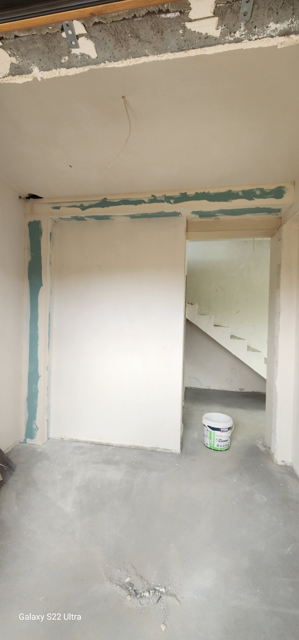 Wnętrze w trakcie prac przygotowawczych do malowania: ściany po szpachlowaniu z widocznymi śladami taśmy malarskiej, surowa podłoga, wiadro z farbą w głębi.