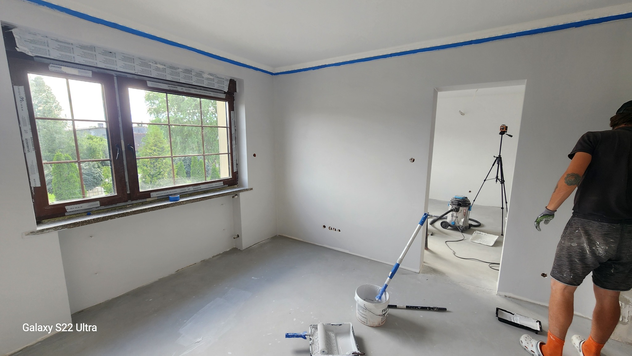 Pokój po malowaniu ścian na szaro, z niebieską taśmą malarską przy suficie i oknach. Widoczne narzędzia malarskie i osoba w trakcie prac wykończeniowych.