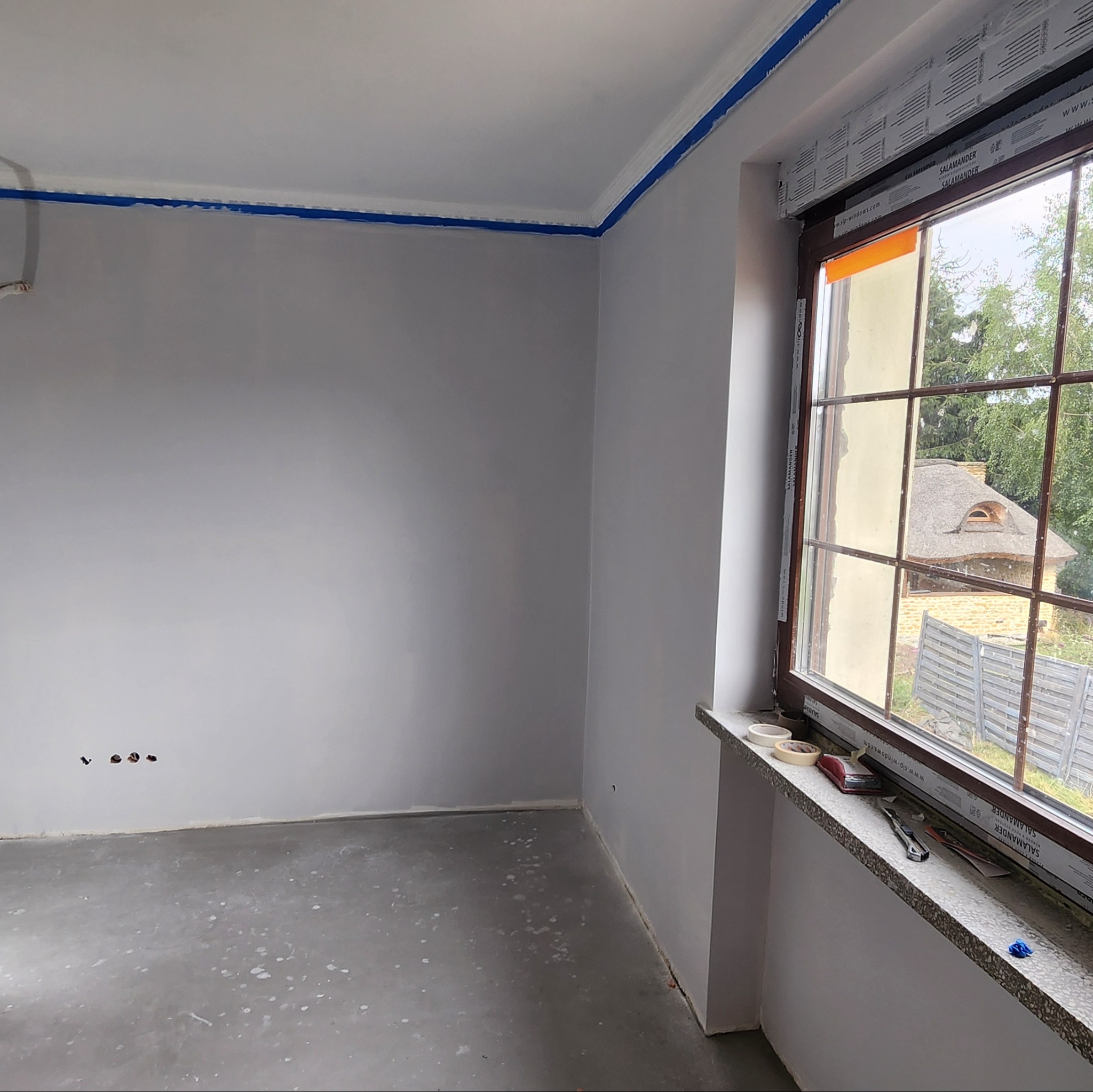 Pokój w trakcie malowania: szare ściany, zabezpieczone taśmą listwy przy suficie i okno. Widoczne narzędzia malarskie na parapecie. W tle budynek z dachem krytym strzechą.