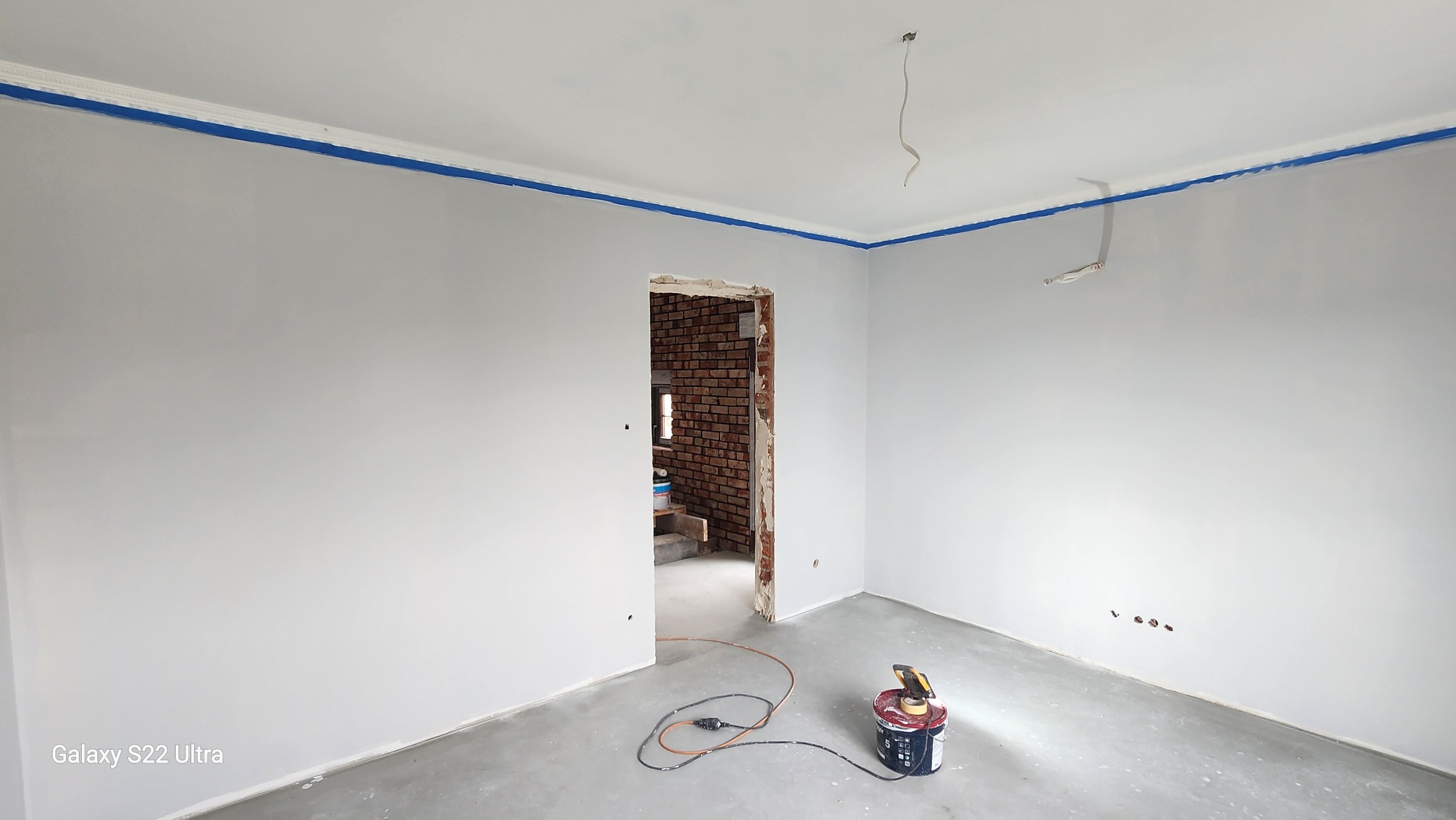 Pokój po malowaniu ścian na szaro, zabezpieczony niebieską taśmą malarską przy suficie. W tle widoczne cegły w przejściu do innego pomieszczenia.