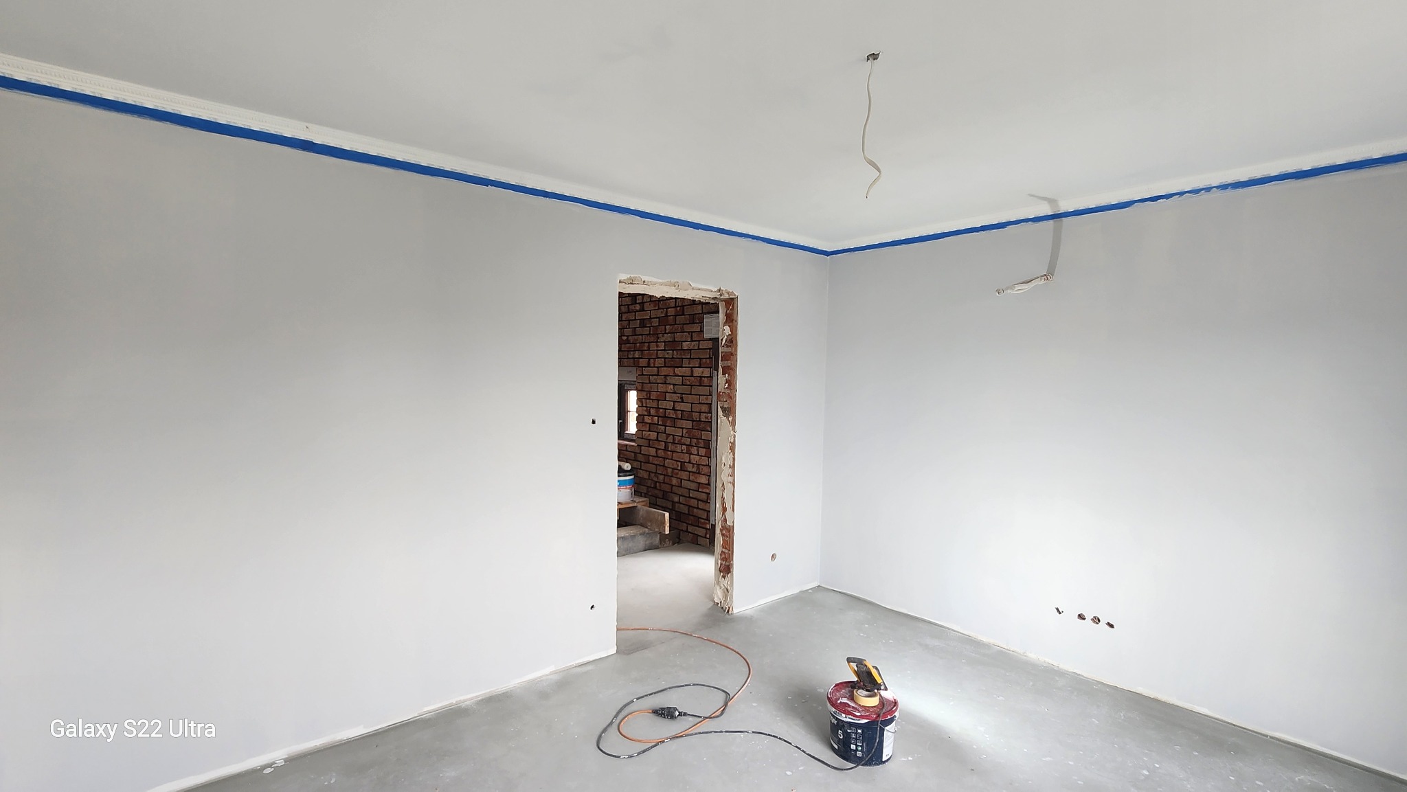 Szare ściany po malowaniu z niebieską taśmą malarską przy suficie, widoczny sprzęt malarski i fragment muru z cegły w przejściu do innego pomieszczenia.