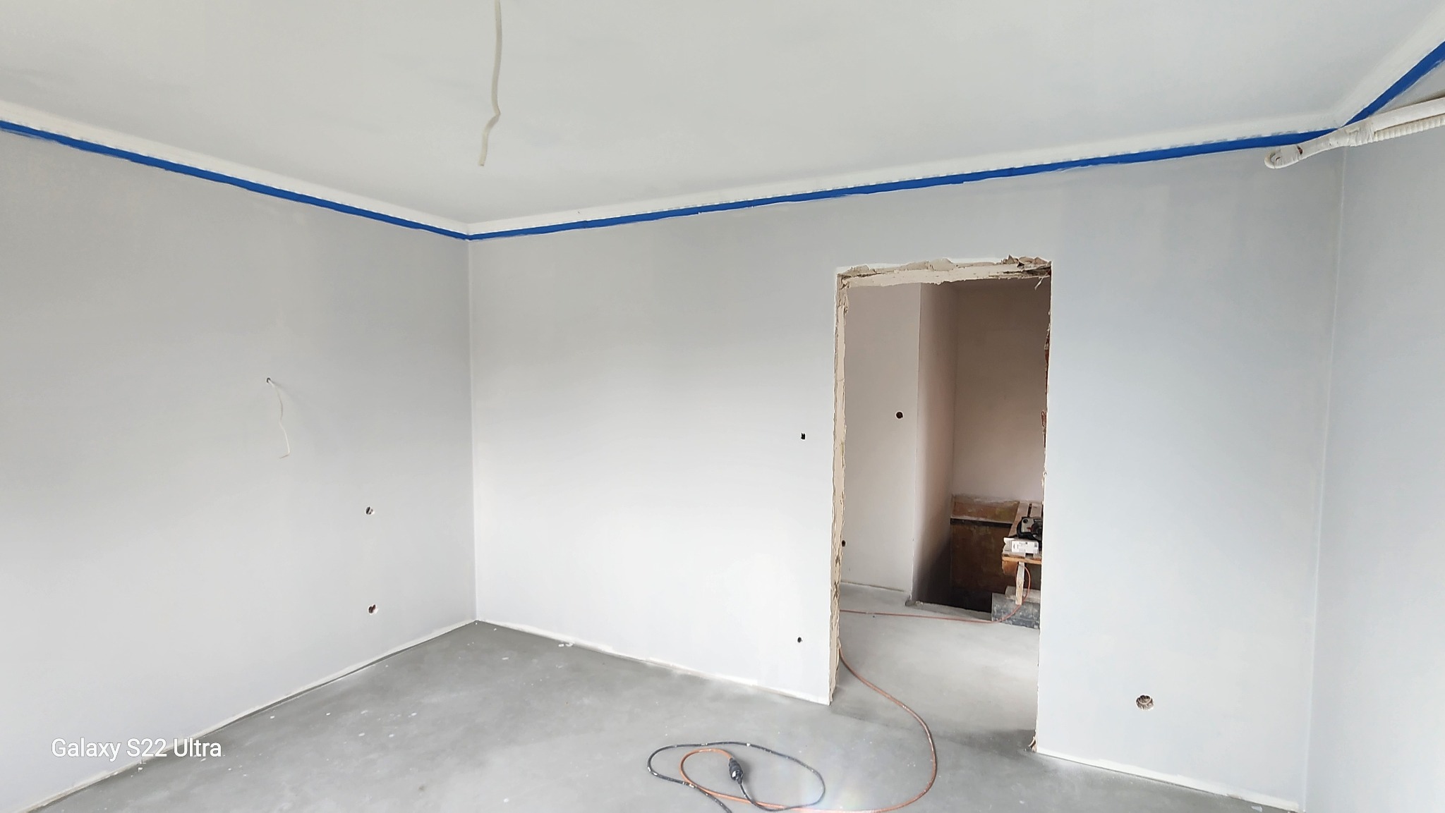 Świeżo pomalowane szare ściany w pokoju z niebieską taśmą malarską przy suficie. Widoczne gniazdka elektryczne i przewody. Podłoga betonowa, surowy stan wykończenia.