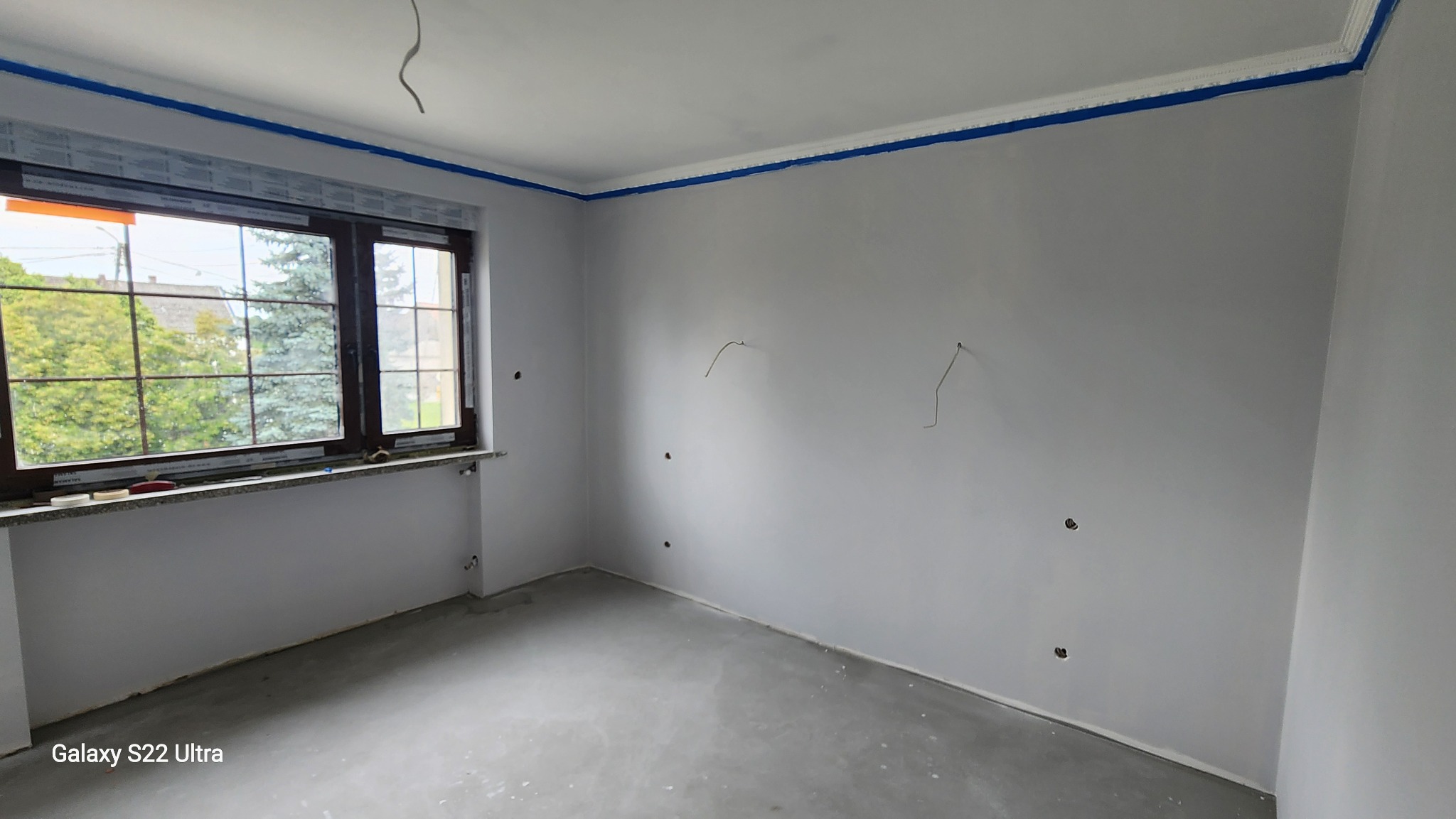 Pokój po malowaniu ścian na szaro, z oknem i listwami przypodłogowymi zabezpieczonymi niebieską taśmą malarską. Widoczne wystające kable elektryczne.